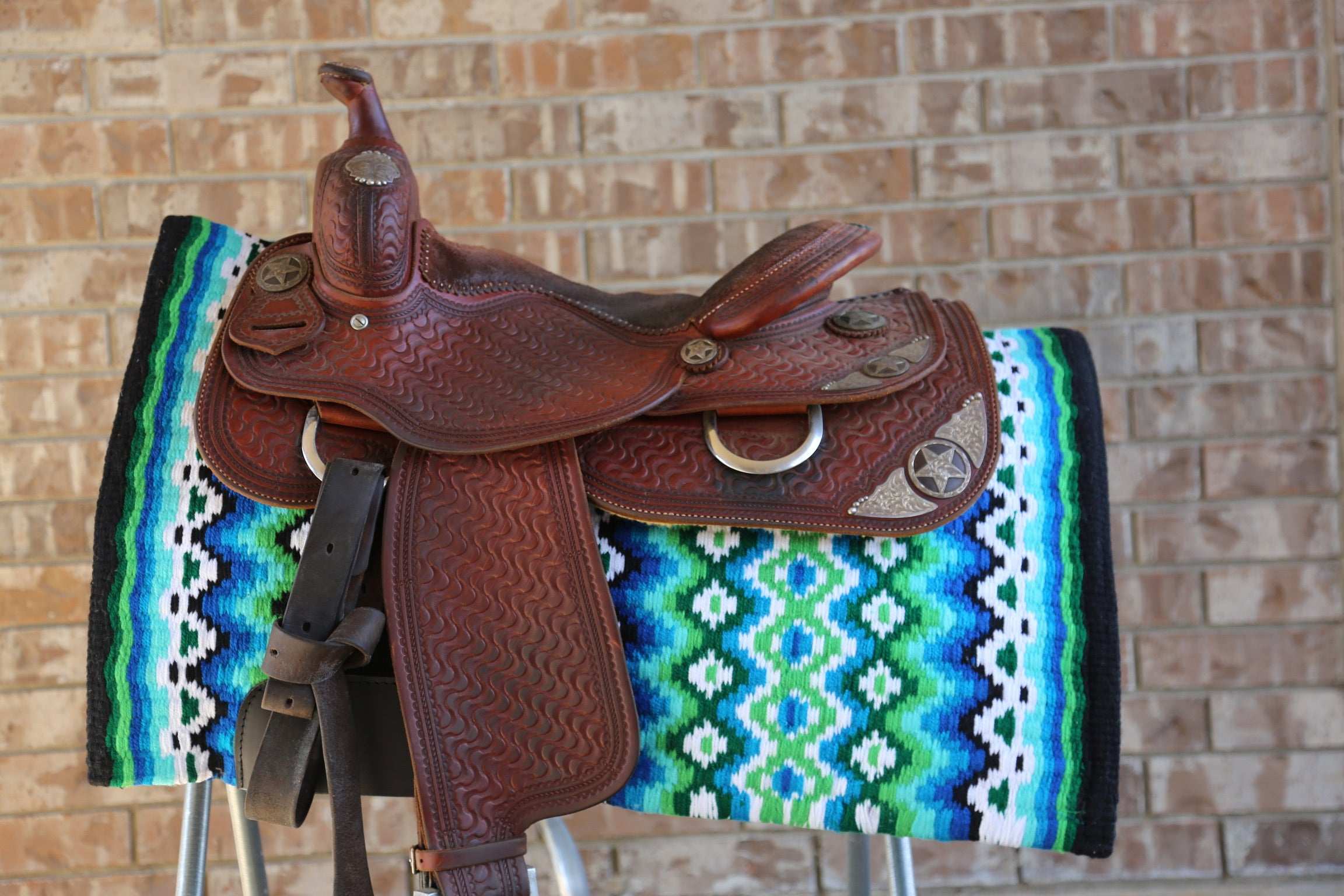 Custom Western Saddle Pad in Blue, Green, White, Black geometric color pattern displayed on a saddle rack and western saddle on top with brick background | Limited Edition
