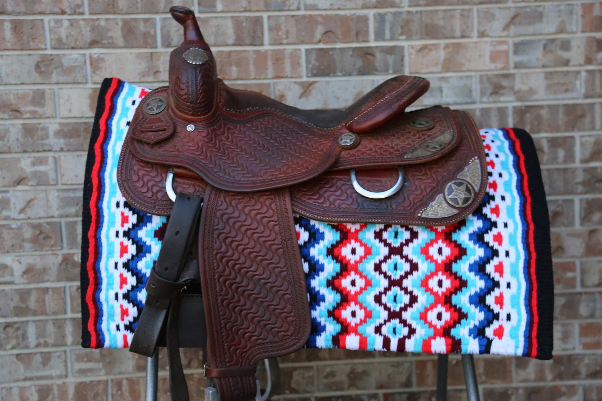 Brown leather saddle on a colorful patterned saddle blanket against a brick wall.