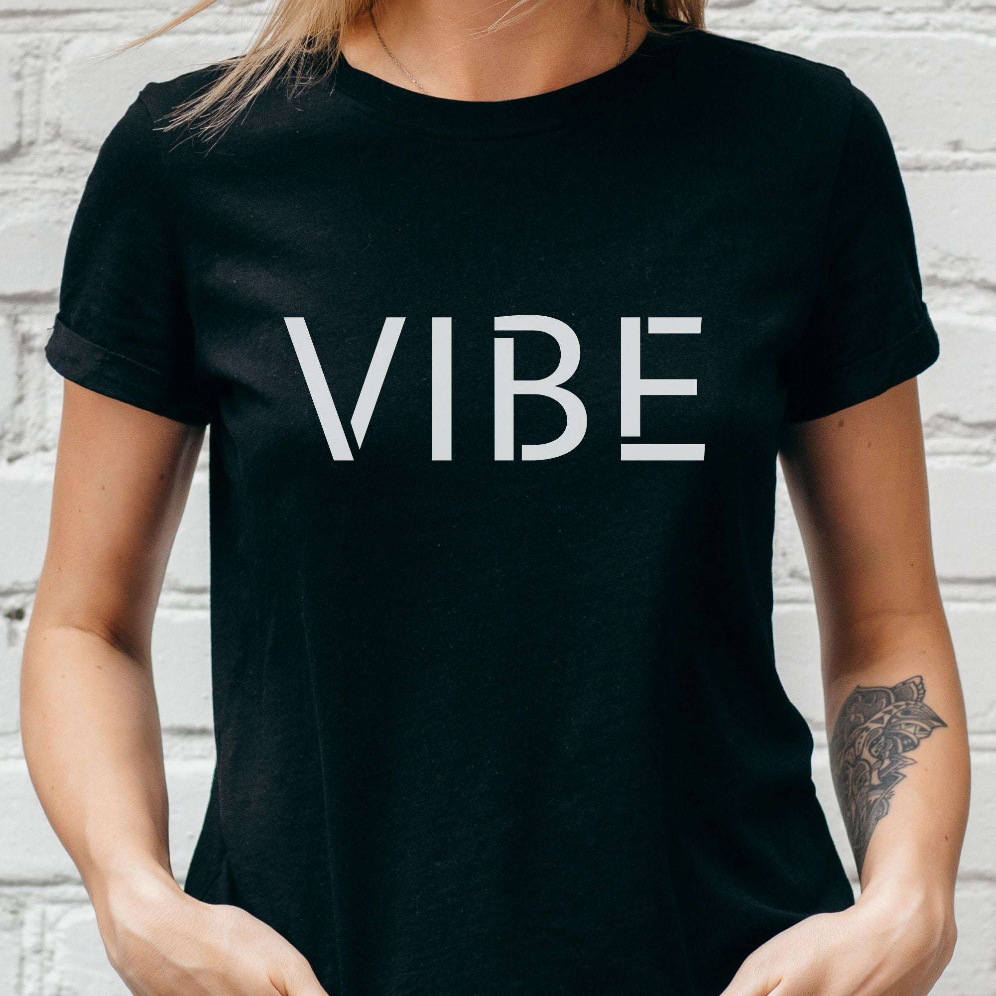 Person wearing a black t-shirt with 'VIBE' printed on it against a white brick wall.